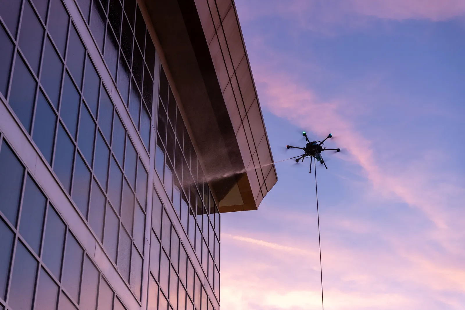 Cleaning drone spraying soft-wash solution across the glass corner of a commercial tower at dusk