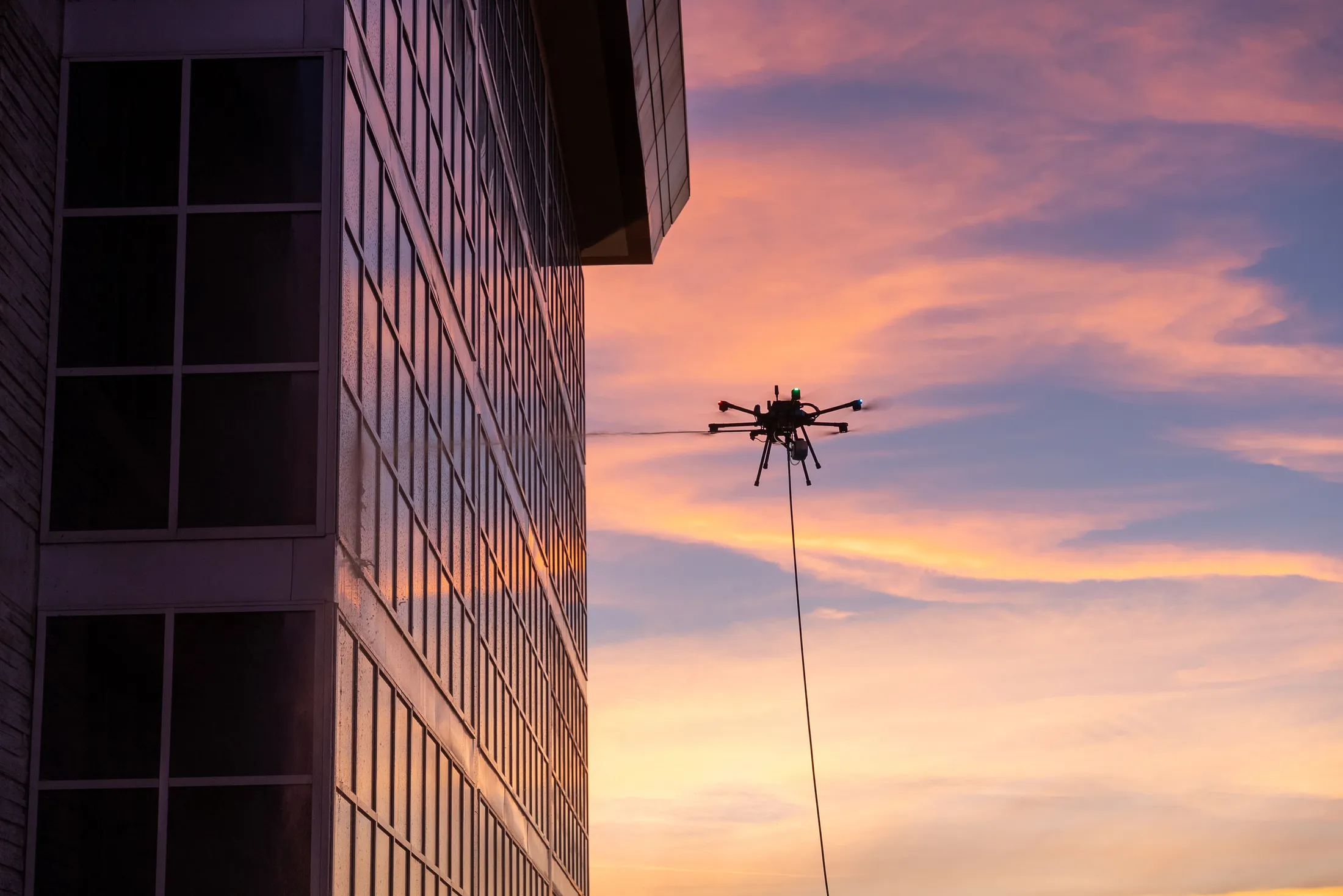 Cleaning drone hovering beside a commercial glass tower during a dusk soft wash