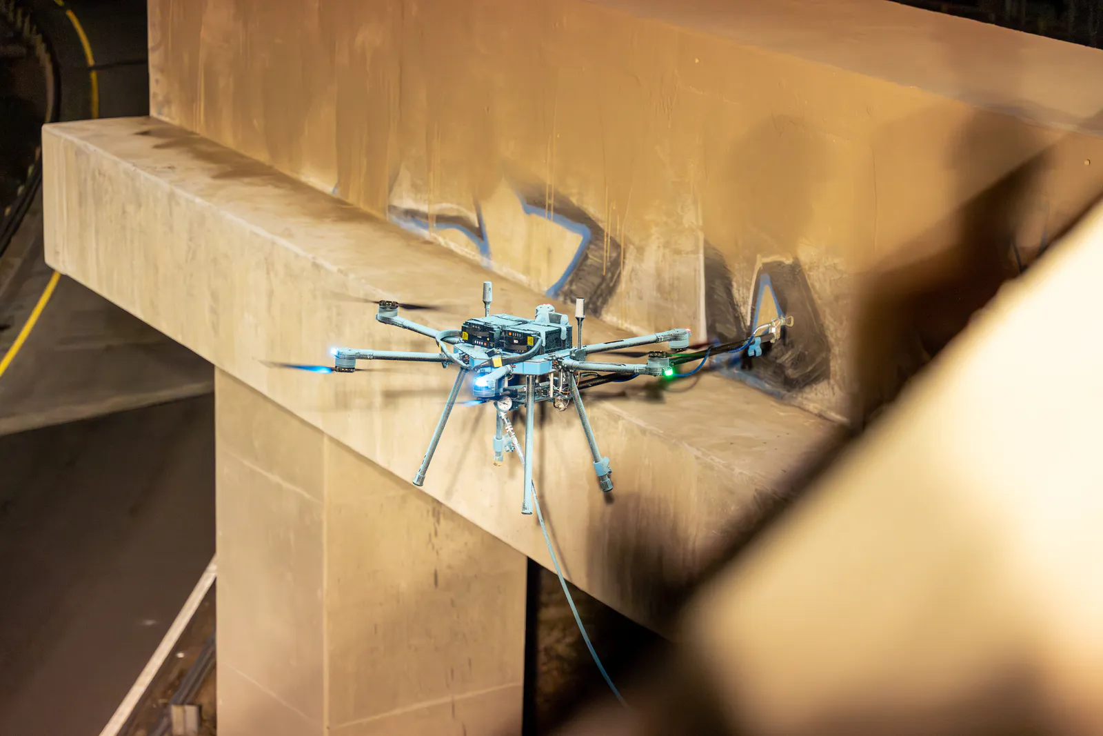 Lucid Bots Sherpa cleaning drone and spray head positioned beside graffiti on a concrete overpass